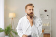 © Studio Romantic - Fixing beard style. Morning preparation of attractive adult man in white bathrobe who fixes his beard looking into camera. Ad of male haircare cosmetics products. Concept of men's care.