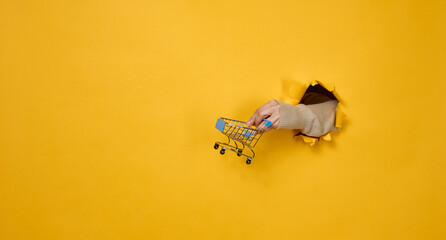 female hand holds a miniature shopping cart on a yellow background, a part of the body sticks out of a torn hole in a paper background. Business concept, start of sales and online shopping