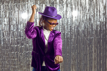 Happy easy going African American woman having fun at a disco party. Cheerful young black girl wearing a purple hat, velvet jacket and party glasses dancing on a shiny silver foil fringe background