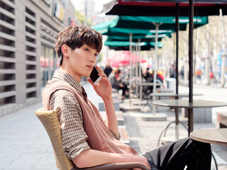 Portrait of handsome Chinese young man with curly black hair in plaid shirt and wool vest sitting outdoor cafe with his mobile phone in hand in sunny day, male fashion, cool Asian young man lifestyle