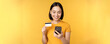 Online shopping. Smiling asian girl using credit card and mobile phone app, paying contactless, order on smartphone application, standing over yellow background