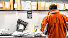 Man Standing At Mailroom Inside Apartment Looking For Packaging. Messenger Deliver Parcel Or Receiver Concept. Guy In Red Uniform Jacket With Pile Of Delivering Bags And Boxes At Distributor Storage.