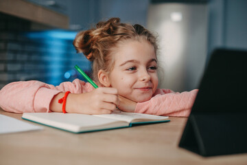 Smiling girl sitting at desk doing homework while doing online educationusing tablet. . Coronavirus school. Quarantine, pandemic. Remote workplace. Smart home.