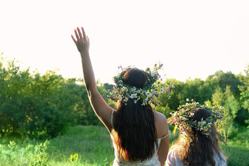 Two girls in flower wreaths on meadow, sunny green natural background. Floral crown, symbol of summer solstice. Slavic ceremony on Midsummer, wiccan Litha sabbat. pagan holiday Ivan Kupala