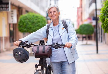 Attractive smiling senior cyclist woman pushing his electric bicycle in a city street looking at camera. Elderly active grandmother enjoying a healthy lifestyle and free time in retirement