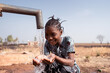 Happy little African girl crouching in front of a waterhole in an arid environment somewhere in the subsaharn region, drinking lots of fresh water from her cupped palms; world water day icon