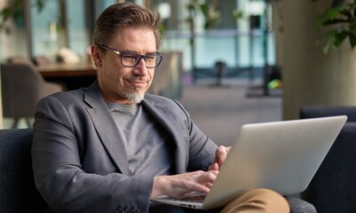 Wall Mural - Businessman using laptop computer in office lobby or on cafe terrace. Happy middle aged man, entrepreneur, small business owner working online.