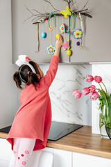 A little dark-haired girl in a pink dress and bunny ears on her head hangs festive easter decor on kitchen cabinet at home.Happy Easter concept.Rear view.