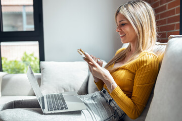 Wall Mural - Beautiful young woman working with her laptop while using mobile phone sitting on a couch at home.