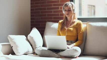 Wall Mural - Video of beautiful young woman working with her laptop while listening to music with earphones sitting on a couch at home.