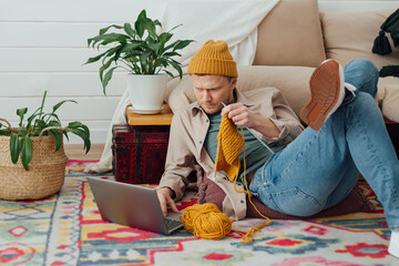 Young serious man looking at laptop. Man learning new hobby, knitting on needles. Knitting project in progress.