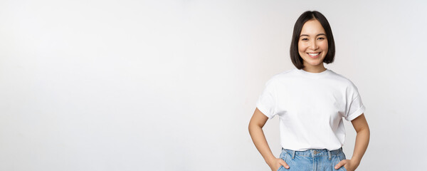 Wall Mural - Lifestyle. Happy modern asian girl, smiling and looking happy at camera, posing in white tshirt and jeans, studio background