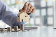 © kamiphotos - Person holding a small wooden house model, stacks of coins on the table, saving money to spend and managing to buy a future home. Money saving ideas to buy a house or residence.