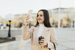 © eduard - Happy smiling beautiful woman holding coffee hot cup and taking photo with smart phone in city while walking in streets.