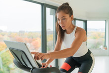 Home Cycle Workout Asian Fit Athlete Woman Training On Stationary Bike Doing Online Cycling Exercise Class Biking During Livestream On Screen.