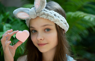 Poster - Portrait of cute little teenage girl enjoying skin care procedures. Beautiful little kid with funny bunny headband cleansing her face by heart shape sponge