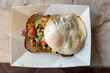 Gourmet Mexican avocado whole wheat toast garnished with poached eggs, lemon zest, red onions, tomatoes, and parsley, on white take-out box and marble table  viewed from the side
