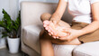 © sorapop - Foot pain, Asian woman sitting on sofa feeling pain in her foot at home, female suffering from feet ache use hand massage relax muscle from soles in home interior, Healthcare and podiatry medical