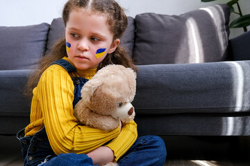 Sign of Ukrainian flag on child's cheek. Ukrainian geopolitics globe crisis. Little girl hold fluffy bear sitting on floor. Sadness longing hope. No war. Hope for the world. Anxiety and anticipation