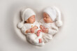 Tiny newborn twin boys in white bodysuits on a white background in white caps. Newborn twins sleep next to their brother on the background of the heart. Two newborn twin boys hugging each other.