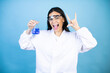 © Irene - Young brunette woman wearing scientist uniform holding test tube over isolated blue background shouting with crazy expression doing rock symbol with hands up