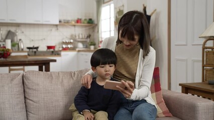 Wall Mural - smiling Asian mother explaining to her preschool son while showing him an educational video on mobile phone in the living room at home