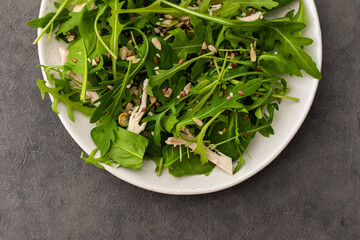 Delicious light salad with arugula and chicken breast sprinkled with various healthy seeds in a plate on a gray background, top view