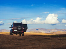 A Black SUV Stands In A Golden Autumn Field Located In The Highlands Under A Blue Sky With Occasional White Cumulus Clouds.