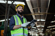 © eakgrungenerd - Portrait of man engineer wear yellow helmet holding tablet and uniform standing at industrial workshop. handsome workman with beard on face ​in factory. Copy space.