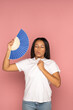 Exhausted african american woman with closed eyes using blue fan suffer from heat sweating, feels sluggish. Black female cooling in hot summer weather, high temperature, isolated on pink background.