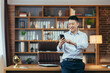 © Liubomir - Successful Asian businessman in modern home office, looks at phone screen, smiles and rejoices, portrait of successful freelancer in shirt, uses phone app