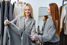 Blonde Affable Caucasian Fashion Consultant Joyfully Helping To Choose Trendy Jacket To Beautiful Redhead Female In Modern Store, Outfits On Hangers In Background. Two Ladies Have Talk, Communication