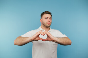 Adult man with bristles and in a white T-shirt stands on a blue background and shows a heart gesture to the camera.