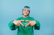 © bodnarphoto - Happy lady with colored hair with a smile on her face shows a heart gesture to the camera on a blue background.
