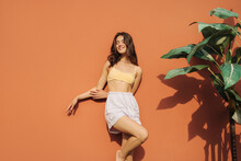 Stylish Young Caucasian Woman Basking In Sun With Her Eyes Closed While Standing Against Red Wall. Brunette Is Wearing Summer Casual Clothes On Warm Day. Season And Leisure Concept.