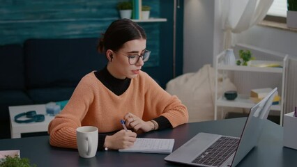 Wall Mural - Female student attending online lesson on videoconference meeting, taking notes on textbook to study university knowledge. Woman using remote teleconference call for learning on laptop.