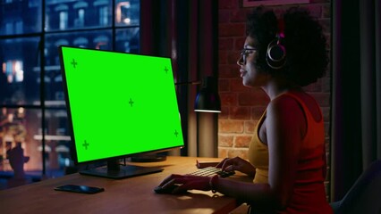 Fototapete - Young Beautiful Female in Headphones Working from Home on Desktop Computer with Green Screen Mock Up Display. Creative woman Checking Social Media, Browsing Internet. Urban City View from Big Window.