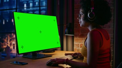 Fototapete - Young Beautiful Female in Headphones Working from Home on Desktop Computer with Green Screen Mock Up Display. Creative woman Checking Social Media, Browsing Internet. Urban City View from Big Window.