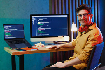 Young fun happy back-end developer software engineer IT specialist programmer man in shirt headhones work at home writing code on laptop pc destop computer analyzing data. Program development concept.