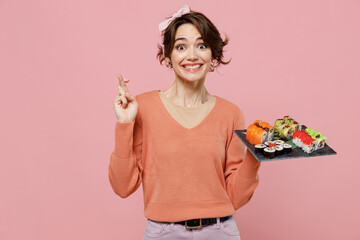 Wall Mural - Young fun woman 20s in casual clothes hold in hand makizushi sushi roll served on black plate traditional japanese food keeping fingers crossed, making wish isolated on plain pastel pink background