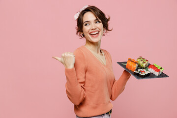 Wall Mural - Young woman in sweater hold in hand makizushi sushi roll served on black plate traditional japanese food point thumb finger aside on workspace area mock up isolated on plain pastel pink background