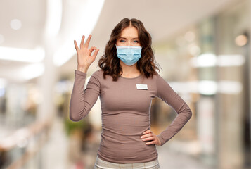 Poster - sale and pandemic concept - happy female shop assistant in medical mask with name tag showing ok gesture over shopping mall background