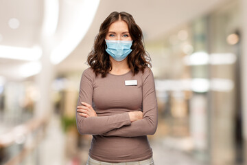 Poster - sale and pandemic concept - happy female shop assistant or saleswoman in medical mask with name tag over shopping mall background