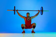 Studio shot of young man in red sportswear exercising with barbell isolated blue background in neon. Sport, weightlifting, power, achievements concept