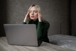 © Екатерина Бучинская - A sad and very tired blonde woman in a green golf is bored working at home with a laptop. Girl sitting at the table in a bad mood in the room. Online learning. Unsightly video conference. High quality