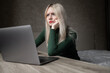 © Екатерина Бучинская - A sad and very tired blonde woman in a green golf is bored working at home with a laptop. Girl sitting at the table in a bad mood in the room. Online learning. Unsightly video conference. High quality