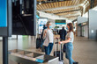 © malajscy - Two travelling women wearing protective masks discussing by flight information board at the airport