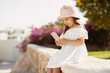© sofiko14 - Little girl in white dress and straw hat using her smartphone, looking at the video, is happy with beautiful smile on background of tropical luxury resort enjoying vacation time, copy space.