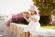 © sofiko14 - Photo of little excited preschool girl holding telephone and waves to mom or friends during video chat wearing white dress and straw hat on the background of tropical city, outside.