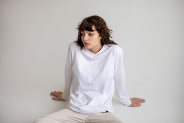 Young woman with curly hair in a white long sleeve t-shirt sits on a white background. Mock-up.